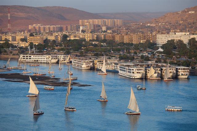Aswan riverfront with feluccas sailing and Nile cruise ships moored below the town