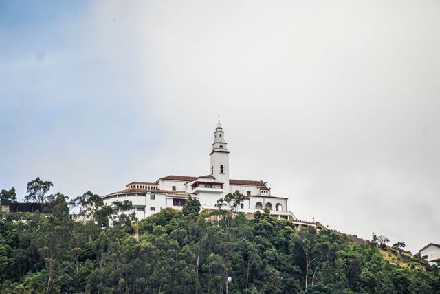 Monserrate Sanctuary's white church and buildings atop a forested hill above Bogota under a cloudy sky