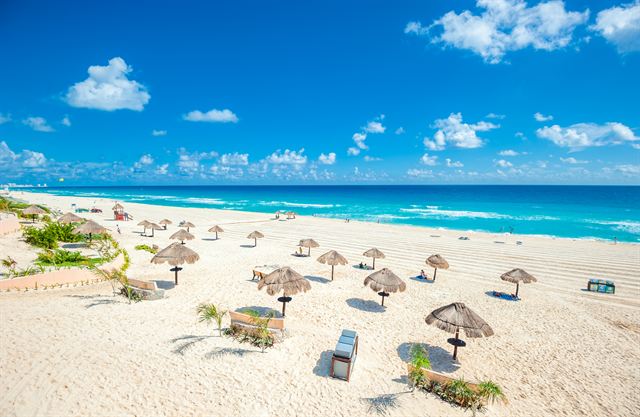 Wide white-sand Cancun beach dotted with thatched palapa umbrellas and loungers under blue sky