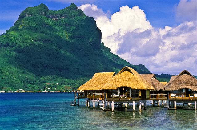 Thatched overwater bungalows on stilts in a turquoise lagoon below a green peak, Fiji