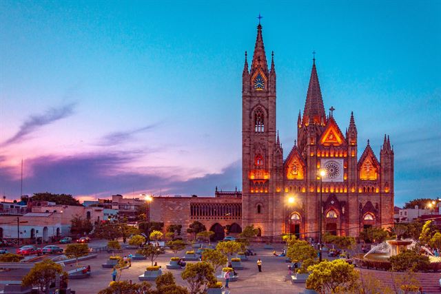 Gothic Templo Expiatorio in Guadalajara lit up at twilight above its tree-lined plaza under a pink sky