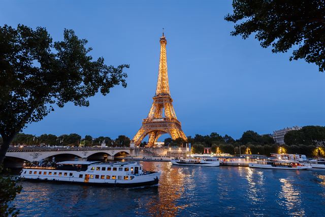 Eiffel Tower lit at dusk above the Seine, with tour boats moored along the river