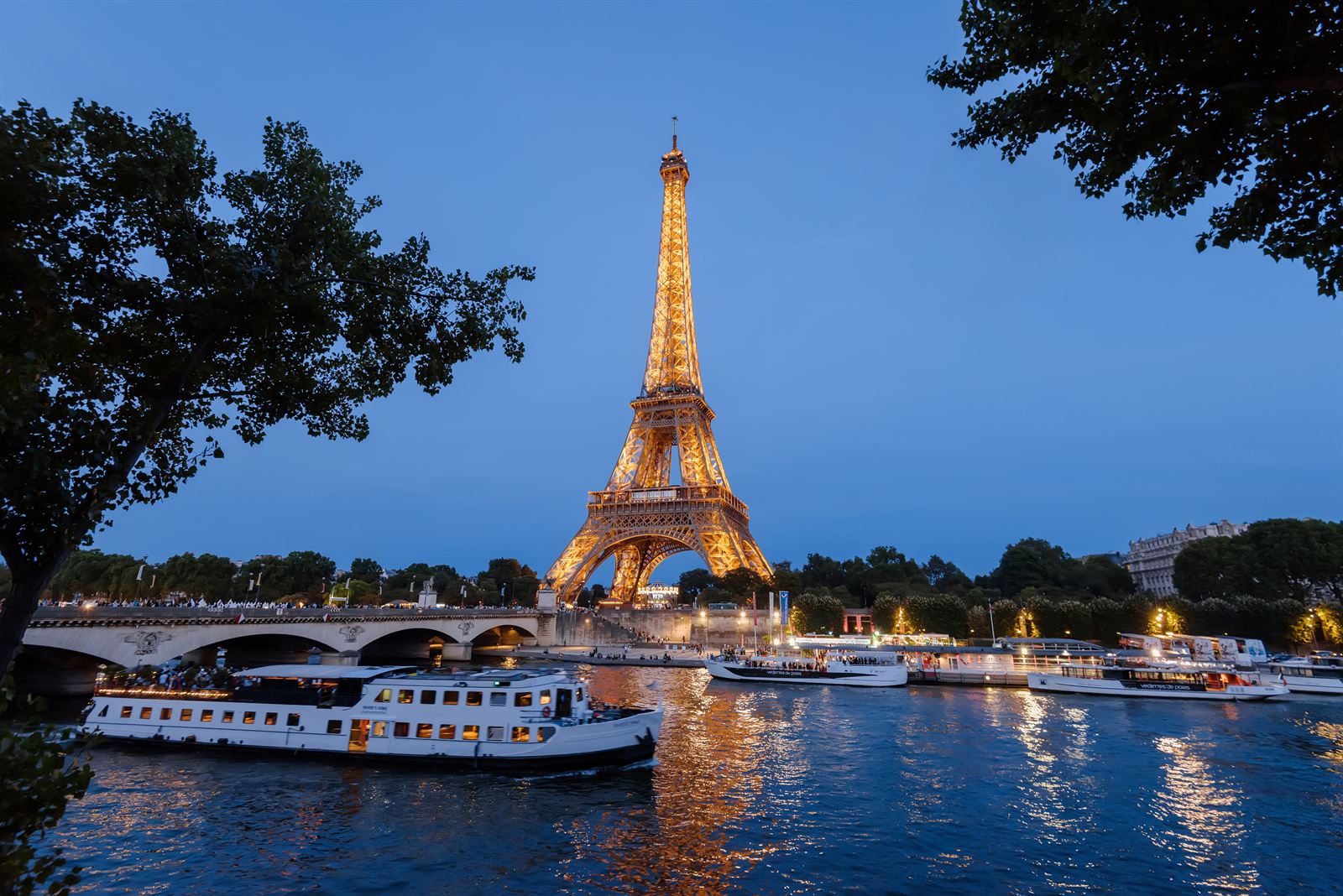Eiffel Tower lit at dusk above the Seine, with tour boats moored along the river