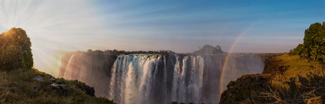 Wide panorama of Victoria Falls with rainbows and sun rays over the gorge